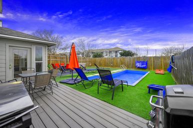 Inviting suburban backyard patio with a small rectangular plunge pool on bright artificial turf, several black lounge chairs and a vibrant orange umbrella, wooden privacy fence, deck dining table and barbecue grill under a vivid blue sky.