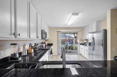Bright suburban galley kitchen with white cabinets, black granite countertops, stainless steel appliances, center sink and faucet, countertop blender and knife block, and sliding glass door opening to a backyard patio with table, chairs and umbrella.