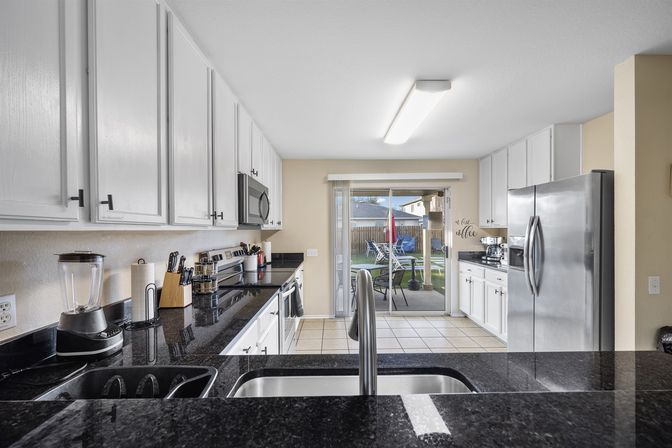 Bright suburban galley kitchen with white cabinets, black granite countertops, stainless steel appliances, center sink and faucet, countertop blender and knife block, and sliding glass door opening to a backyard patio with table, chairs and umbrella.