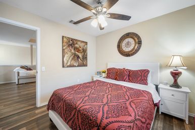 Bright modern bedroom with red patterned bedding, white headboard, bedside lamp and round wall clock, ceiling fan, wood floors and view into adjacent living area