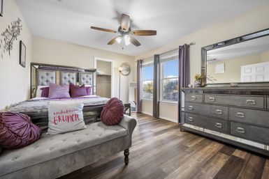 Cozy sunlit master bedroom with tufted gray headboard, purple accent pillows and round bolsters, a tufted bench with a playful “weekends are for BRUNCH” cushion, wood ceiling fan, double windows with gray curtains, mirrored dresser and wood-look floors.