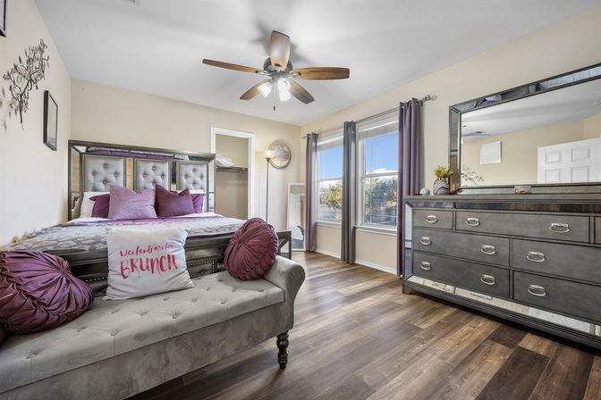 Cozy sunlit master bedroom with tufted gray headboard, purple accent pillows and round bolsters, a tufted bench with a playful “weekends are for BRUNCH” cushion, wood ceiling fan, double windows with gray curtains, mirrored dresser and wood-look floors.