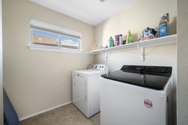 Compact bright laundry room with white top-load washer and matching dryer, wall shelf holding detergent bottles, fabric softener and an iron, tiled floor and a small horizontal window.