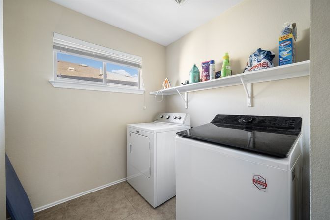 Compact bright laundry room with white top-load washer and matching dryer, wall shelf holding detergent bottles, fabric softener and an iron, tiled floor and a small horizontal window.