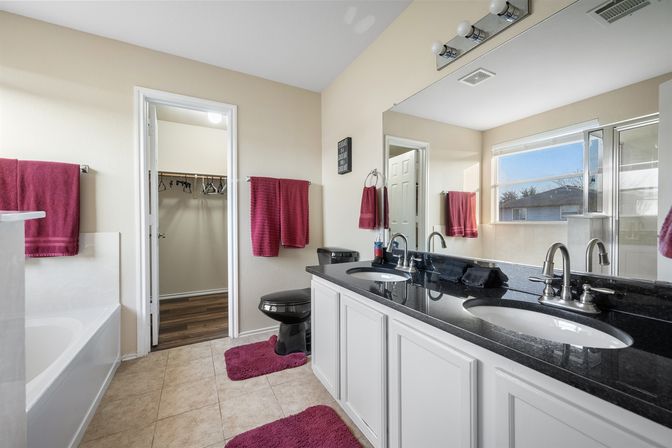Bright suburban master bathroom with black granite double vanity and twin chrome sinks, large mirror, soaking tub, black toilet, magenta towels and rugs, beige tile floor, and open walk-in closet visible.