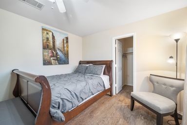 Bright modern bedroom interior with wooden sleigh bed and gray comforter, hardwood floors, tufted gray accent chair, floor lamp, canal painting on the wall and an open closet doorway.