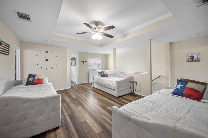 Bright open loft with three white tufted daybeds, Texas flag accent pillows, wood-look flooring, ceiling fan, and oversized wall clock