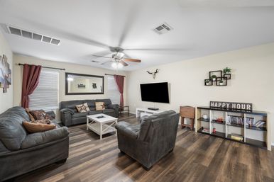 Cozy living room with dark leather sofas and armchair around a white coffee table on wood-look floors, wall-mounted flat-screen TV, ceiling fan, red curtains, large mirror and decorative shelving with a 'Happy Hour' sign.