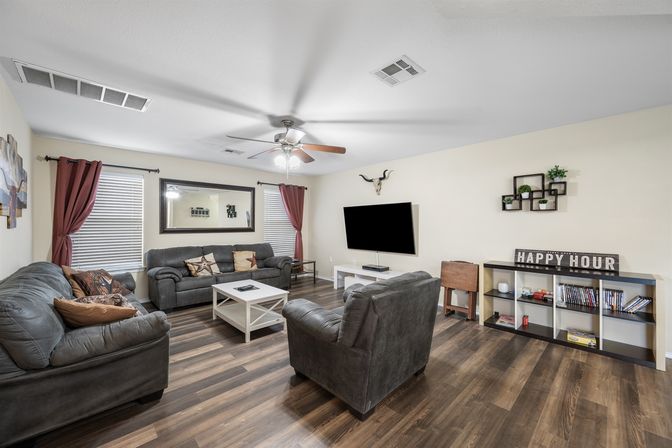 Cozy living room with dark leather sofas and armchair around a white coffee table on wood-look floors, wall-mounted flat-screen TV, ceiling fan, red curtains, large mirror and decorative shelving with a 'Happy Hour' sign.