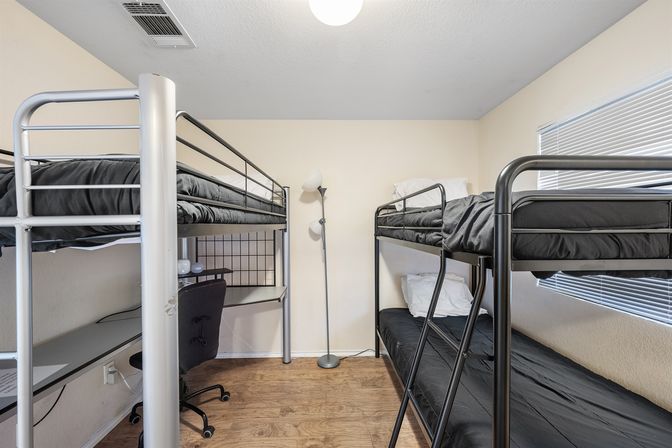 Compact bedroom with two black metal bunk beds facing each other, black bedding, a lofted desk and rolling chair under one bed, floor lamp, window blinds, and wood-look flooring