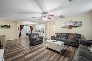 Cozy open-concept living room and dining area with dark wood-look floors, gray sectional and armchairs, white coffee table, ceiling fan, longhorn wall art, and a kitchen with red-curtained dining nook.
