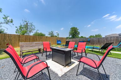 Inviting suburban backyard patio with a square fire pit surrounded by red chairs, a wooden bench and lounge chairs on gravel and turf, fenced yard under a clear blue sky.