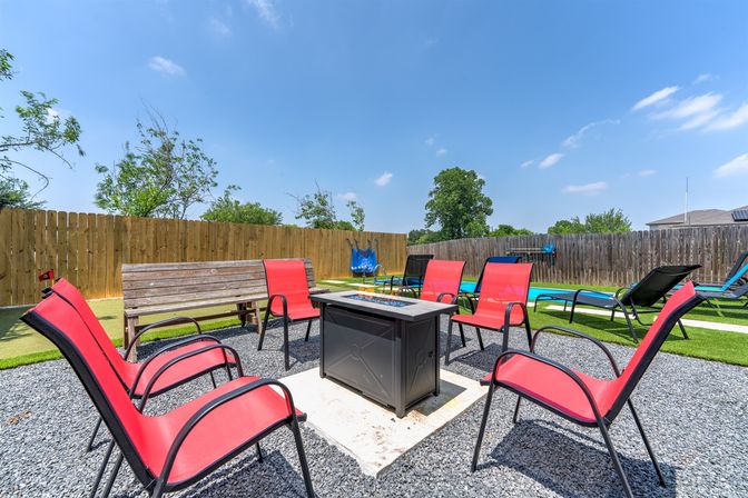 Inviting suburban backyard patio with a square fire pit surrounded by red chairs, a wooden bench and lounge chairs on gravel and turf, fenced yard under a clear blue sky.