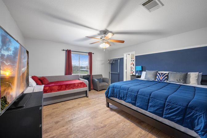 Cozy modern master bedroom with navy accent wall, king bed in blue comforter, gray daybed with red throw, wall TV showing a sunset, armchair, ceiling fan and wood floors.