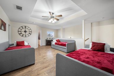 Bright upstairs loft bedroom with three gray daybeds topped with red throws, wood plank floors, ceiling fan, large wall clock and a dresser under the window.