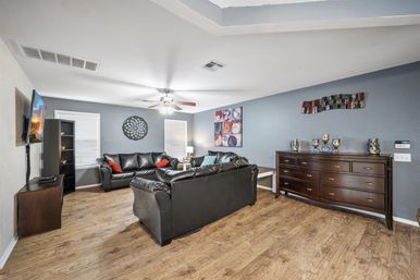 Modern living room interior with gray-blue walls, wood-look flooring, black leather sofas, ceiling fan, mounted TV, colorful wall art and a dark wood dresser.