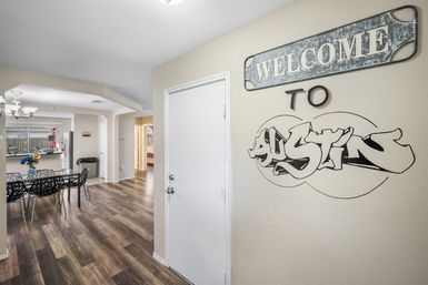 Bright modern home entryway with a rustic "WELCOME" sign and graffiti-style 'Austin' wall art, white door and wood-look floors opening to an open dining area with glass table, black chairs, vase of flowers and stainless steel kitchen appliances.