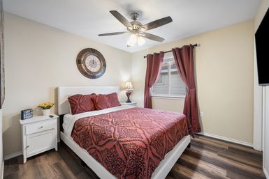 Cozy bedroom interior with bold red patterned bedding on a white platform bed, matching pillows, round wall clock above the headboard, white nightstands with lamp and flowers, ceiling fan, red curtains and wood-look flooring.
