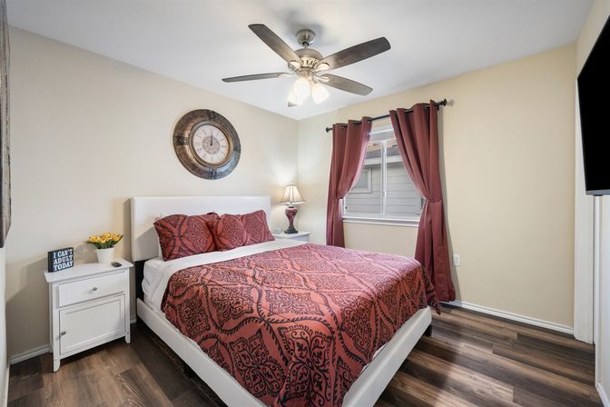 Cozy bedroom interior with bold red patterned bedding on a white platform bed, matching pillows, round wall clock above the headboard, white nightstands with lamp and flowers, ceiling fan, red curtains and wood-look flooring.