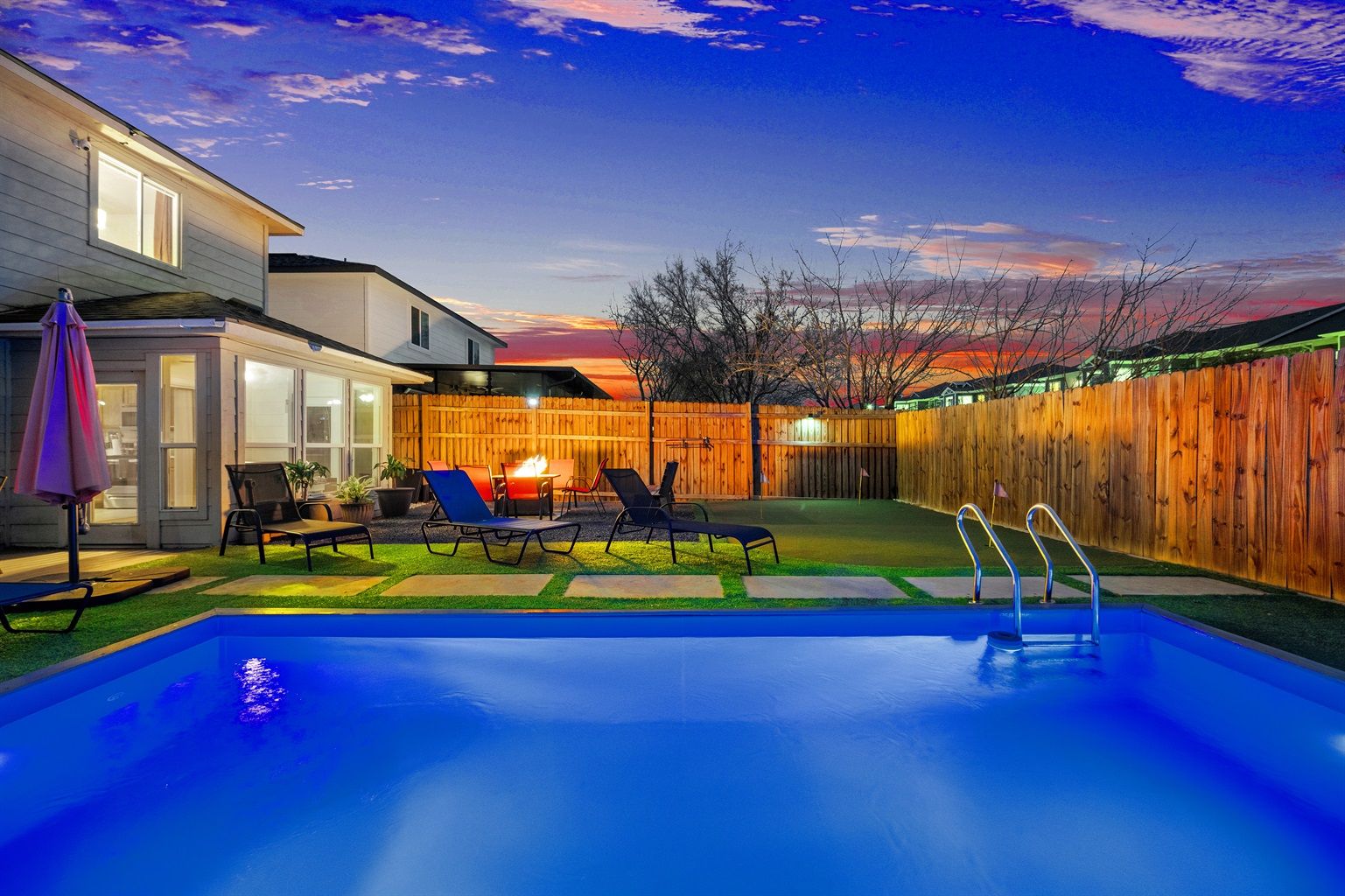Suburban backyard pool at sunset — blue-lit rectangular pool with metal ladder, lounge chairs and umbrella, glowing fire pit on a patio, wooden privacy fence and two-story house under a colorful twilight sky.