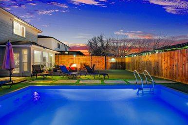 Suburban backyard pool at sunset — blue-lit rectangular pool with metal ladder, lounge chairs and umbrella, glowing fire pit on a patio, wooden privacy fence and two-story house under a colorful twilight sky.