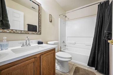 Bright modern residential bathroom with white tub-shower and chrome fixtures, oak vanity with white countertop, framed mirror and black shower curtain