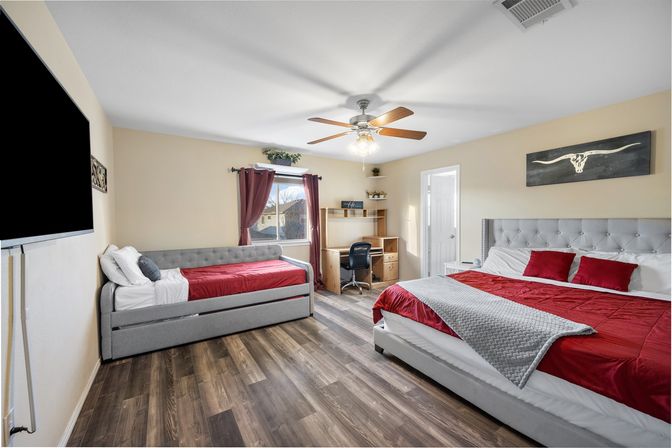 Bright modern master bedroom with two gray upholstered beds (daybed with trundle and king), red bedding and gray throws, wood-look flooring, ceiling fan, wall-mounted TV, desk by a window with burgundy curtains, and longhorn wall art.