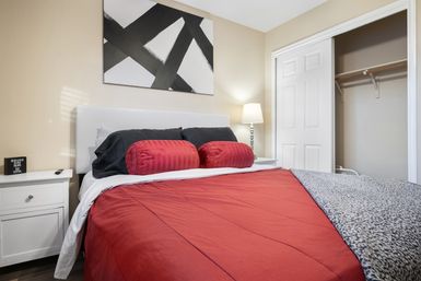 Cozy modern apartment bedroom with red comforter and matching bolster pillows, white upholstered headboard, black accent pillows, abstract black-and-white wall art, white nightstand with lamp, and an open closet.