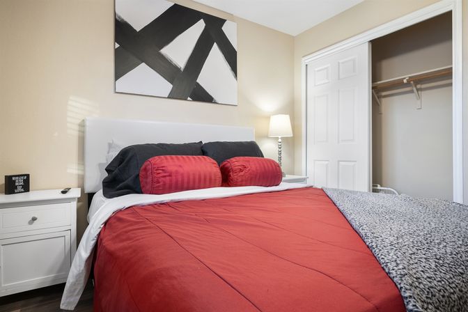 Cozy modern apartment bedroom with red comforter and matching bolster pillows, white upholstered headboard, black accent pillows, abstract black-and-white wall art, white nightstand with lamp, and an open closet.