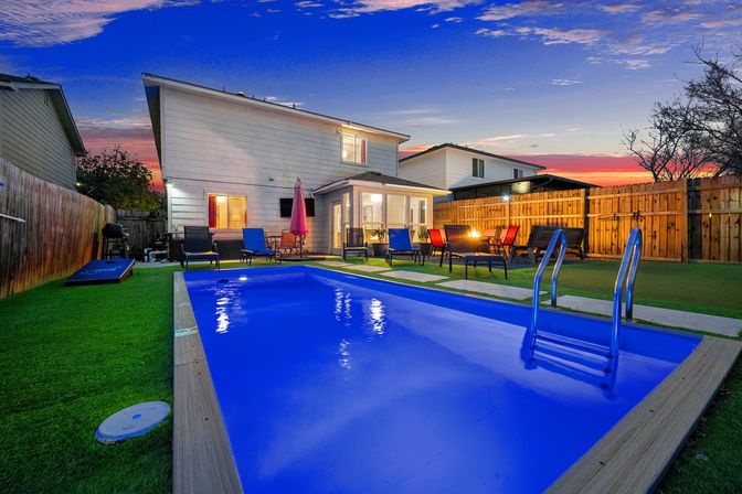 Glowing blue backyard pool at sunset in a fenced suburban yard, with metal ladder, lounge chairs, umbrella and cozy patio seating