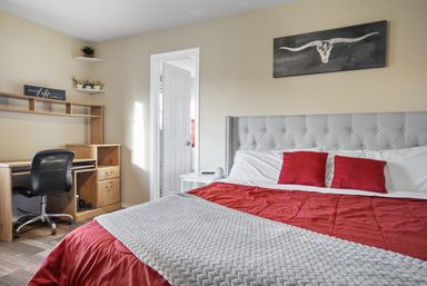 Bright cozy bedroom with gray tufted headboard, red bedding and pillows, textured gray throw, longhorn wall art, and a wooden desk home-office nook with a black swivel chair by an open doorway.