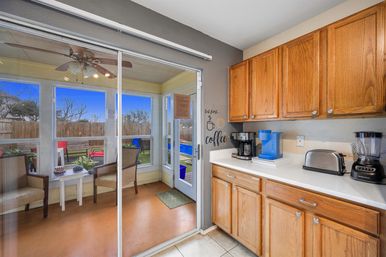 Sunny kitchen coffee station with oak cabinets and white countertop holding a drip coffee maker, blue single-serve brewer, toaster and blender; sliding glass door opens to an enclosed sunroom with ceiling fan, chairs and a view of a fenced backyard patio.