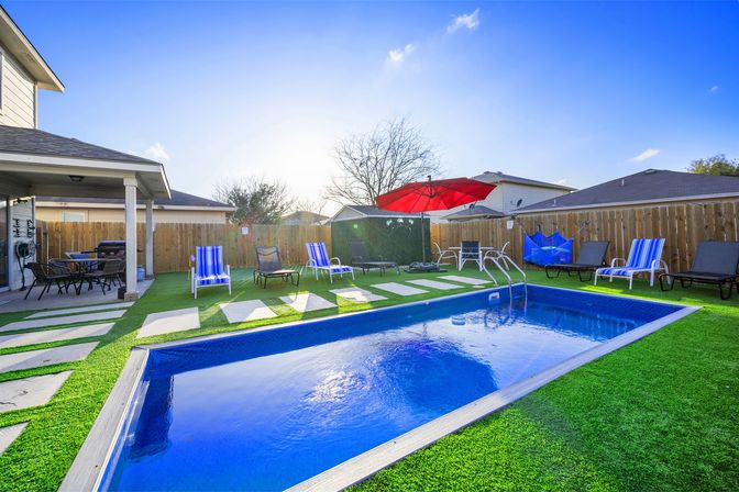 Sunny suburban backyard with rectangular blue in-ground pool, green artificial turf, striped lounge chairs, red patio umbrella, covered dining patio and wooden privacy fence