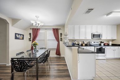Bright open-concept kitchen and dining area with glass-top table, black leaf-pattern chairs, sunflower centerpiece, white cabinets and stainless appliances, burgundy curtains and mixed wood and tile floors.