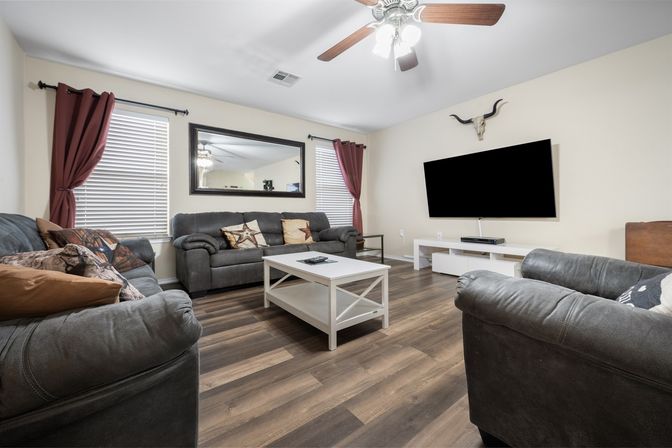 Cozy modern living room with dark gray sofas and star-pattern pillows, white coffee table on wood-look floors, wall-mounted flat-screen TV, large mirror between two windows with red curtains, ceiling fan and decorative longhorn skull.