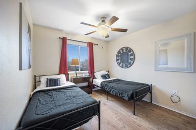 Cozy bright guest bedroom with two black metal twin beds and black bedding, wood floors, ceiling fan, nightstand with lamp beneath a window framed by red curtains, plus a large round wall clock and mirror.