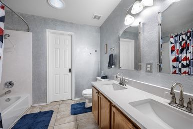 Bright residential bathroom with double sink vanity and chrome faucets, white countertop over wood cabinets, large mirror and lights, toilet and tub-shower with red‑blue geometric curtain, gray textured walls and blue bath mats on tiled floor.