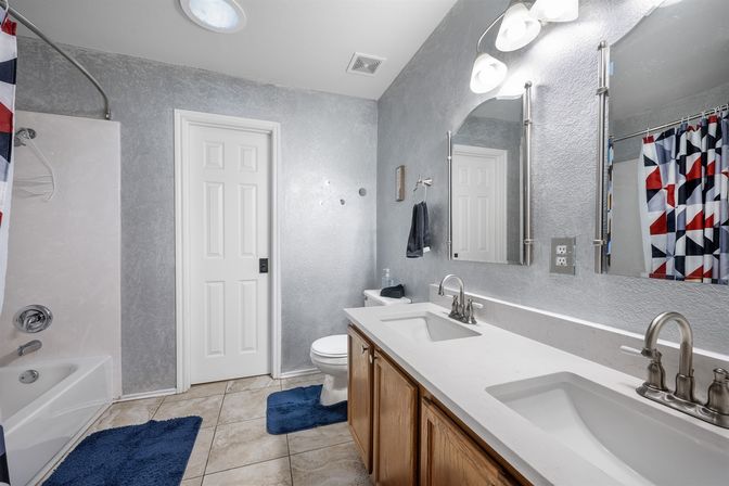 Bright residential bathroom with double sink vanity and chrome faucets, white countertop over wood cabinets, large mirror and lights, toilet and tub-shower with red‑blue geometric curtain, gray textured walls and blue bath mats on tiled floor.