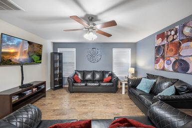 Cozy modern living room interior with black leather sofas, hardwood floor, ceiling fan, wall-mounted TV, colorful wall art and decorative throw pillows.