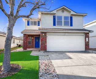 Curb view of a two-story suburban home with white siding and brick porch, bright blue front door and shutters, attached two-car garage, leafless front tree, rock-lined driveway and green lawn.