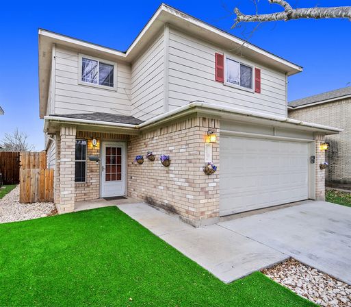 Two-story suburban house with beige brick and light siding, attached two-car garage, concrete driveway, bright green lawn, hanging flower baskets and warm porch lights.