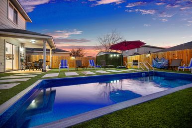 Evening suburban backyard with a glowing rectangular pool at sunset, striped lounge chairs and red patio umbrella, covered patio dining area, stepping-stone turf and wooden privacy fence.