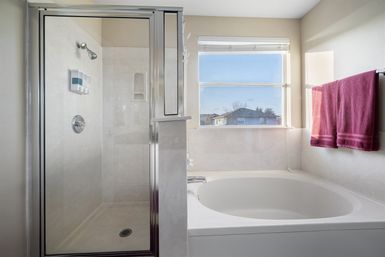 Sunlit home bathroom with glass-enclosed shower, large oval soaking tub under a window with neighborhood view, and magenta towels on a rack.