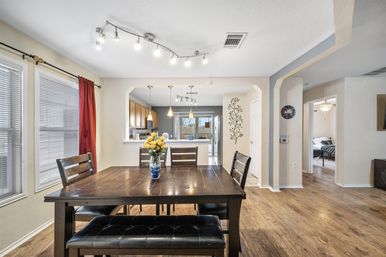 Bright open-concept dining room in a suburban home — dark wood table with bench seating and vase of yellow flowers, hardwood floors, large windows with red curtain, and view into a modern kitchen with pendant lights and stainless refrigerator.