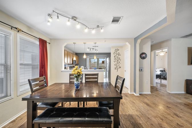 Bright open-concept dining room in a suburban home — dark wood table with bench seating and vase of yellow flowers, hardwood floors, large windows with red curtain, and view into a modern kitchen with pendant lights and stainless refrigerator.