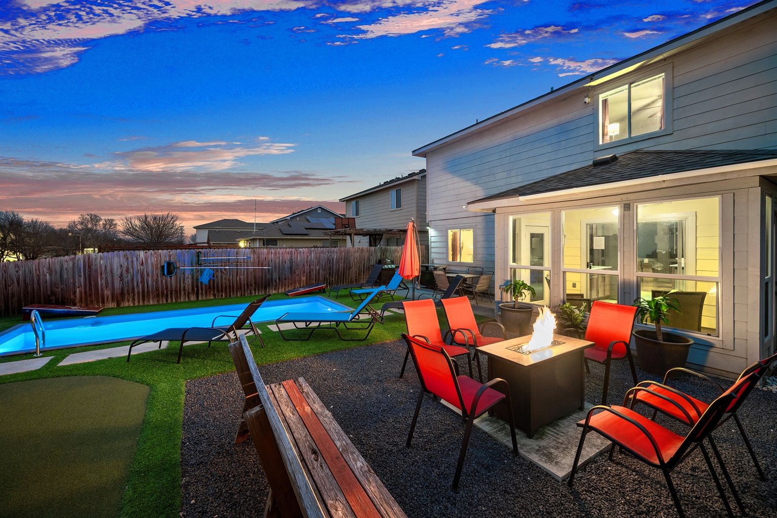 Inviting suburban backyard patio at dusk with a lit rectangular pool, red-cushioned chairs around a glowing fire pit, lounge chairs, umbrella, and a two-story house.