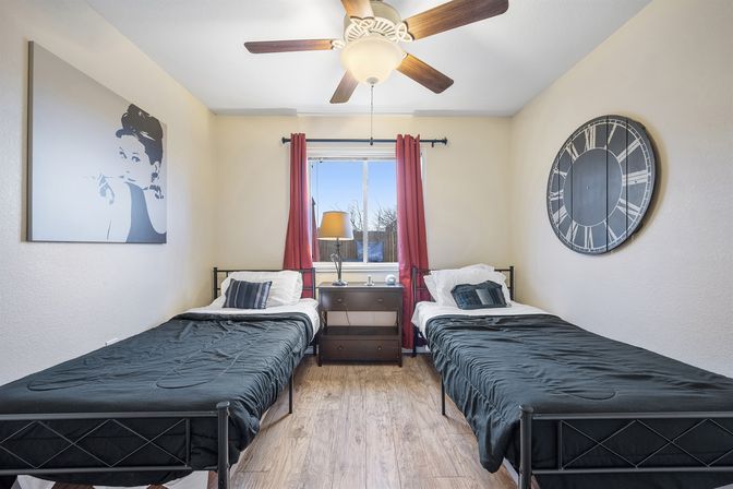 Cozy twin bedroom with matching black metal beds and dark bedding, wood-look floor, central nightstand and lamp beneath a window with red curtains, ceiling fan, oversized round wall clock and monochrome portrait.