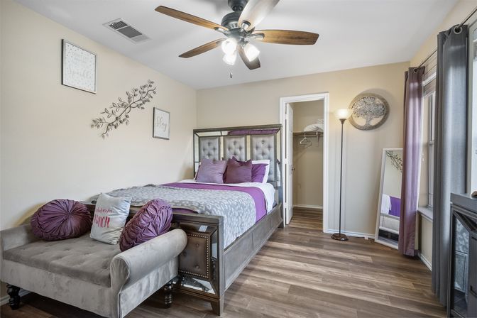 Cozy modern bedroom with tufted headboard and purple bedding, gray chaise at the foot, wood-look floors, ceiling fan, floor lamp, full-length mirror, wall art and open closet.