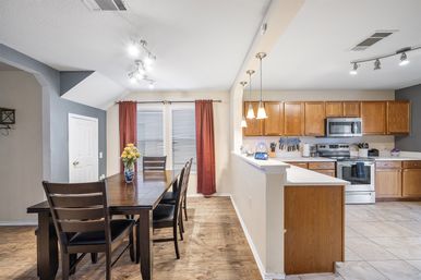 Bright open-concept kitchen and dining area with a dark wood table and chairs, vase of flowers, red curtains, pendant lights, oak cabinets and stainless-steel appliances.