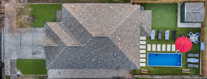 Aerial drone view of a suburban home showing the roof, driveway and landscaped backyard with a rectangular blue swimming pool, red patio umbrella, lounge chairs, stepping-stone path and green turf.